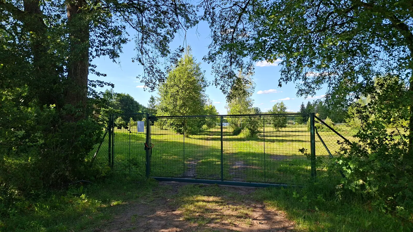 Zdjęcie w orientacji poziomej ukazuje metalową, zieloną bramę wjazdową ustawioną w środku kadru, która oddziela leśną ścieżkę od słonecznej, trawiastej polany. Brama wykonana jest z siatki drucianej i stanowi główny punkt centralny kompozycji, tworząc naturalne przejście w głąb zielonego terenu. Po obu stronach bramy rozciąga się bujna roślinność, w tym duże drzewa liściaste, których korony rzucają cień na pierwszy plan, tworząc naturalną ramę dla całego obrazu. W oddali widać błękitne niebo z delikatnymi chmurami oraz rozproszone drzewa porastające łąkę. Na lewym skrzydle bramy zawieszona jest niewielka, biała tabliczka z tekstem, której treść jest nieczytelna z tej odległości. Całość sceny jest oświetlona jasnym światłem słonecznym, co podkreśla głębię terenu i sielski, spokojny charakter tego miejsca.